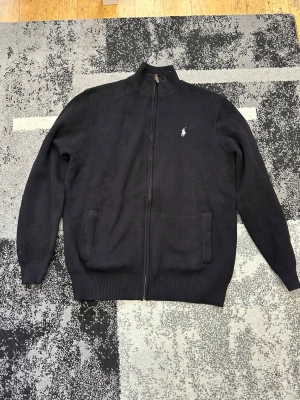 Svart ziptröja från Polo Ralph Lauren - Klassisk svart ziptröja från Polo Ralph Lauren med hög krage och vit broderad logga på bröstet. Tröjan har två fickor framtill, ribbade muddar och är tillverkad i ett mjukt stickat material. Perfekt för en clean och stilren look.