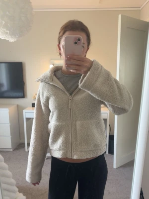 Beige teddyjacka med dragkedja - Mysig beige teddyjacka med dragkedja framtill och två stora fickor. Jackan har en hög krage och är gjord i fluffigt, mjukt material som ger en avslappnad och trendig look. Perfekt för kyliga dagar och enkel att matcha med olika outfits.