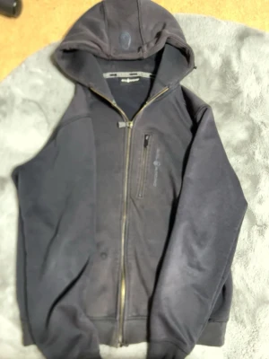 Svart hoodie från Sail Racing - Säljer en svart hoodie från Sail Racing med dragkedja framtill och huva. Hoodien har en diskret logga på bröstet och på huvan, samt fickor på sidorna. Tillverkad i mjukt och bekvämt material, perfekt för en avslappnad stil.