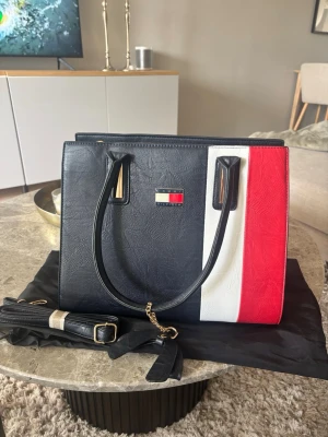Handväska i färgblock från Tommy Hilfiger - Snygg handväska från Tommy Hilfiger i svart, vitt och rött färgblocksmönster. Väskan har två handtag, avtagbar axelrem med gulddetaljer och klassisk logga framtill. Tillverkad i konstläder med struktur. Perfekt för dig som gillar stilrena accessoarer. Ingen aning ifall äkta köpt i Spanien där av priset😁