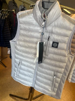 Ljusgrå dunväst från Stone Island - Snygg ljusgrå dunväst från Stone Island med hög krage och dragkedja framtill. Västen har två sidofickor med dragkedja och en bröstficka. Klassisk Stone Island-logga på bröstet. Perfekt för lager-på-lager och streetwear-stil.