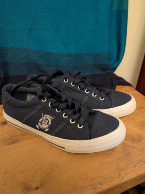 Marinblå sneakers Vinson Polo Club - Snygga marinblå sneakers från Vinson Polo Club med vita sulor och matchande marinblå skosnören. Skorna har en rund tå, metallöljetter och ett broderat märke på sidan. Tillverkade i textilmaterial för en avslappnad och sportig look.