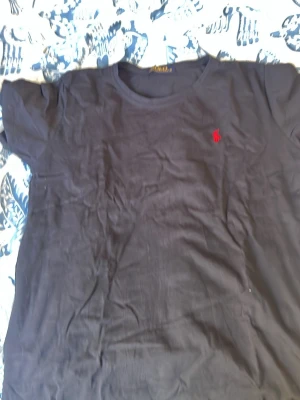 Svart t-shirt från Polo Ralph Lauren - Svart t-shirt från Polo Ralph Lauren med klassisk passform och korta ärmar. T-shirten har en liten röd broderad logga på bröstet och är tillverkad i mjuk bomull. Perfekt till jeans eller shorts.