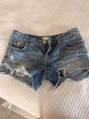 Blå jeansshorts från Gina Tricot - Säljer ett par blå jeansshorts från Gina Tricot i storlek 158. Shortsen har råa, fransiga kanter och slitna detaljer framtill. Klassisk femficksmodell med knapp och dragkedja. Perfekta för varma dagar!