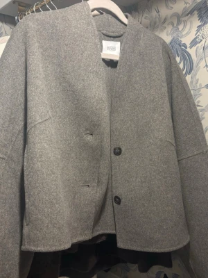 Grå ulljacka med knappar - Stilren grå jacka i ull med två svarta knappar framtill. Modellen är rak och något boxig med lång ärm och enkel design utan krage. Perfekt för dig som gillar minimalistisk och clean stil. Från & Other Stories i London och finns ej i denna grå färg i Sverige. Aldrig använd!
