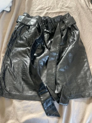 Svart kortkjol i skinnimitation - Snygg svart kortkjol i skinnimitation med croco-mönster och bredt knytband i midjan. Kjolen har en papperspåse-midja som ger en trendig look och passar perfekt till en cool outfit.