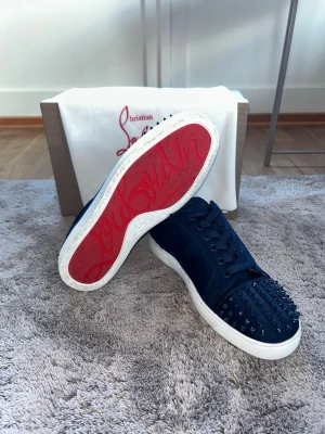 Blåa loubs med spikar - Exklusiva Christian Louboutin-sneakers 👟🔥 Stilrena svarta sneakers med den ikoniska röda sulan och ett exklusivt detaljerat uttryck. De dekorativa nitarna framtill ger skorna en lyxig look som verkligen sticker ut. Perfekta för dig som vill kombinera streetwear med en elegant och exklusiv känsla