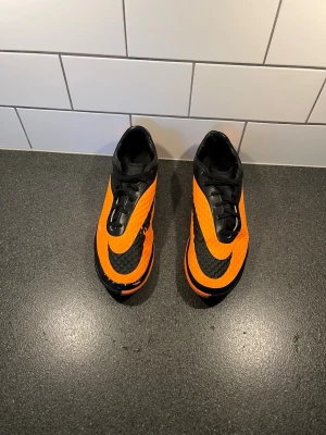 Orange och svarta Nike fotbollsskor - Säljer ett par Hypervenom futsal skor från Nike i orange och svart. Skorna har en modern design med stora Nike-loggor och mönstrad ovansida för bättre bollkontroll. Yttersulan är orange med greppvänligt mönster, perfekt för spel inomhus. Jag säljer dessa skor eftersom som att jag inte kör futsal, jag kör fotboll på hög nivå, så jag vill bara få bort skorna.