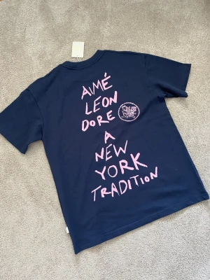 Mörkblå Aimé Leon Dore t-shirt - Snygg mörkblå t-shirt från Aimé Leon Dore med rosa tryck både fram och bak. Framsidan har ett hjärta och texten 'Aimé', medan baksidan har ett stort grafiskt tryck med texten 'A New York Tradition'. Klassisk passform och rund hals i mjuk bomull.