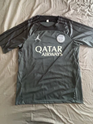 PSG Svart/grå träningströja - Paris Saint-Germain-träningströjan har korta ärmar och rund hals, samt ett vitt Jordan-märke på framsidan. Tillverkad i ett lätt och ventilerande material, perfekt för fotbollsträning.