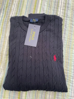 Mörkblå kabelstickad tröja Polo - Kabelstickad mörkblå tröja från Polo Ralph Lauren med rund halsringning och den klassiska röda logotypen broderad på bröstet. Tröjan har ribbade muddar och är tillverkad i mjukt material som passar perfekt till lager på lager.