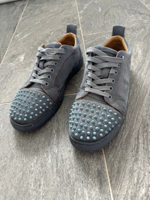 Grå sneakers med nitar och röd sula - Unika grå sneakers i mocka med ljusblå nitar på tån och klassisk röd sula. Skorna har grå snörning och en låg silhuett. Insidan är fodrad i beige läder för extra komfort. Perfekta för dig som vill sticka ut med din stil.