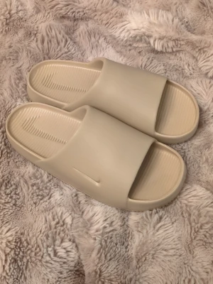 Beige calm slides från Nike - Hej! Säljer dessa så dom inte passade som jag trodde. Tyvärr har jag inte boxen eller kvitto då min hund paja båda två. Endast testade. Nästintill nyskick Säljer ett par stilrena beige slides från Nike. De har en clean design med bred rem över foten och diskret Nike-logga på sidan och i sulan. Tillverkade i mjukt och bekvämt syntetmaterial med räfflad innersula för extra komfort.
