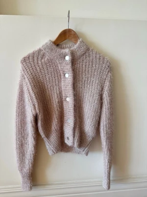 Ljusrosa fluffig kofta med knappar - Supermjuk och fluffig ljusrosa kofta med lång ärm och högt ribbat krage. Koftan har stora vita knappar framtill och ett grovt stickat mönster som ger en mysig känsla. Perfekt för lager på lager och chill dagar.
