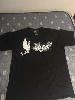 Svart v-lone t-shirt med vit tryck på ryggen som ett V - Säljer en svart t-shirt med vit grafik framtill, där det står 'LONE' i ett rökigt typsnitt och en stor fågel syns bredvid texten. T-shirten har rund hals och korta ärmar, perfekt för en streetwear-look.