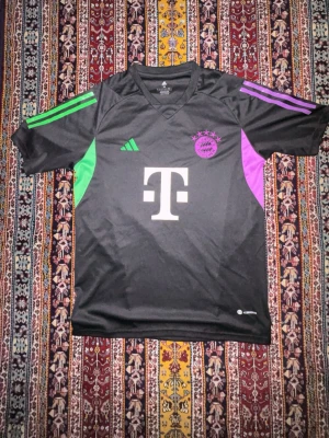 Svart Bayern München matchtröja - Snygg svart matchtröja från Adidas för FC Bayern München med gröna och lila detaljer. Tröjan har V-ringning, korta ärmar och klubbmärke samt Adidas-logga på bröstet. Tillverkad i lätt och ventilerande material, perfekt för fotboll eller träning.