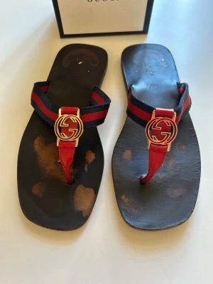 Äkta Gucci sandaler  - Snygga sandaler från Gucci med klassisk röd och svart rem i tyg och guccis ikoniska guldfärgade GG-logga framtill. Sandalerna har en tårem i rött läder och svart lädersula. Köpta på Gucci Stockholm. Dem är i perfekt skick. Sulan kan smörjas in med skocreme så att slitaget på sulans färg inte blir tydlig. Perfekta för en trendig och avslappnad look.