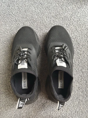 Svarta Steve Madden sneakers - Snygga svarta sneakers från Steve Madden med mesh-ovandel och detaljer i syntet. Skorna har svarta elastiska snören, vit logotyp på plösen och dragflik bak. Perfekta för en trendig och sportig look.