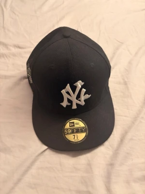 Svart New Era 59FIFTY NY keps - Snygg svart New Era 59FIFTY keps med klassiskt New York Yankees broderat vitt logotyp framtill och World Series-märke på sidan. Platt skärm och guldfärgad storleksdekal kvar på skärmen. Perfekt för dig som gillar streetwear och sportig stil.