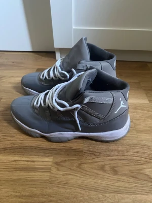 Gråa Air Jordan 11 sneakers - Snygga gråa Air Jordan 11 sneakers med vita detaljer och vita skosnören. Skorna har en blank finish på vissa partier och en tydlig Jumpman-logga på sidan. Sulan är vit med grå inslag och ovandelen är i syntetmaterial och textil.