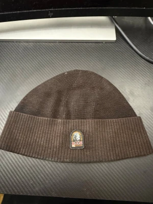 Mörkbrun ribbstickad mössa - Snygg mörkbrun mössa med bred ribbad uppvikt kant och ett litet märke framtill. Klassisk modell som sitter skönt på huvudet och passar perfekt till höst och vinter. Materialet ser ut att vara mjukt och stretchigt, troligen bomull eller akryl.