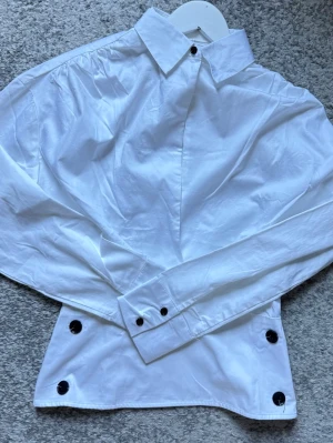 Deep Neck Overlap Shirt från NA-KD - Stilren vit skjorta i bomull med klassisk krage och svarta knappar vid hals, ärmslut och nederkant. Skjortan har lång ärm och en loose passform som ger en avslappnad look. Perfekt för dig som gillar minimalistisk stil.