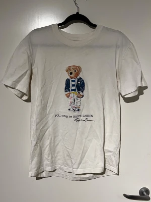 Vit Polo Bear t-shirt från Ralph Lauren - Vit t-shirt med Polo Bear-tryck från Ralph Lauren. T-shirten har en klassisk passform och korta ärmar. Trycket visar en björn i blå jacka och randig tröja. Perfekt för dig som gillar streetwear och ikoniska prints.