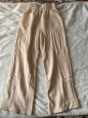 Beige mjukisbyxor med raka ben - Säljer ett par beige mjukisbyxor med raka ben och hög midja. Byxorna har resår i midjan med dragsko och två sidofickor. Tillverkade i mjukt och bekvämt material, perfekta för chill dagar. Passar dig som gillar en avslappnad och enkel stil.