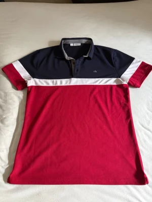 Calvin Klein Golf Piké - Snygg pikétröja från Calvin Klein Golf med marinblå överdel, vit rand och röd nederdel. Pikén har aldrig använts. Kragen har trycket 'CALVIN KLEIN GOLF' på insidan och tröjan har korta ärmar samt knappar vid halsen. Perfekt för en sportig och stilren look. 100% polyester 
