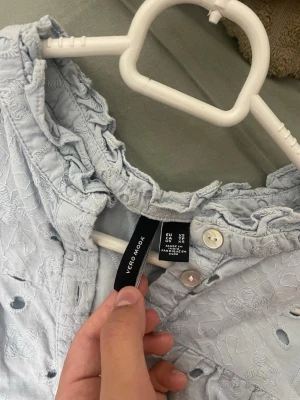 Ljusblå broderad blus Vero Moda - Supersöt ljusblå blus från Vero Moda med broderade detaljer och hålmönster. Blusen har volang vid kragen, knappar framtill och långa ärmar. Perfekt för en fräsch och trendig look till våren eller sommaren.