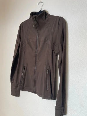 Brun sportig jacka med dragkedja st 4 - Snygg brun jacka med hög krage och hel dragkedja framtill. Jackan har två fickor med dragkedja och en figurnära passform. Perfekt för dig som gillar en sportig och stilren look.