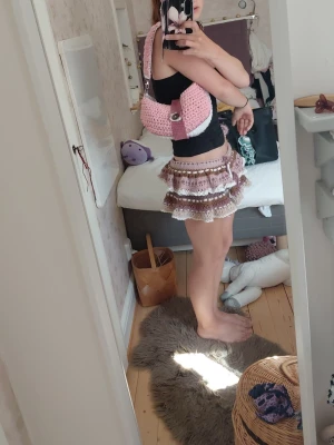 Rosa virkad axelväska - Säljer en superfin handvirkad axelväska i rosa och vitt med bred axelrem och dekorativ knapp framtill. Väskan har en mjuk, chunky känsla och stängs med ett band över locket. Perfekt för dig som gillar unika accessoarer och vill sticka ut.