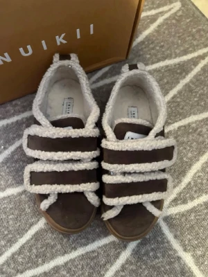 inukki skor - Unika bruna sneakers från Inuikii med tre breda kardborreband och fluffig ullkant runt hela skon. Ovandelen är i mocka och sulan är i gummi. 