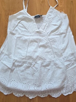 Vit broderad linnetopp med spetskant - Söt vit linnetopp med tunna axelband och v-ringad hals. Toppen har broderade detaljer och spetskant nertill som ger en romantisk känsla. Perfekt för varma dagar och sommarens alla äventyr.