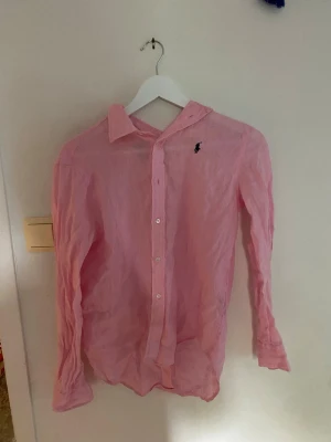 Rosa linneskjorta från Polo Ralph Lauren - Säljer en klassisk rosa skjorta från Polo Ralph Lauren i svalt linne. Skjortan har lång ärm, knappar framtill och den ikoniska broderade loggan på bröstet. Perfekt för en stilren och avslappnad look.