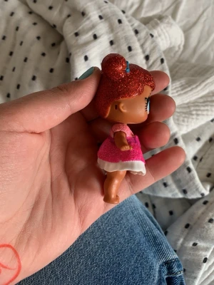 Liten docka med glitterhår - Söt liten plastdocka med rött glitterhår uppsatt i en boll och stor blå ögon. Dockan har en rosa klänning med vit kant och rosa axelband. Perfekt som leksak eller samlarfigur.