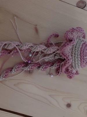 Virkad manet håraccessoar rosa/beige - Unik handgjord håraccessoar i form av en manet, virkad i mjukt garn i rosa och beige toner. Dekorerad med små pärlor på tentaklerna för extra detalj. Perfekt att fästa i håret eller på väskan för en kreativ touch.
