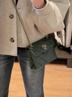 Mörkgrön quiltad axelremsväska - ÄR BORTREST MEN SKA LÄGGA UT FLER BILDER. Snygg mörkgrön handväska med quiltat mönster och silverfärgad kedjerem. Väskan har ett dekorativt spänne framtill och kompakt, boxig form. Perfekt för dig som vill ha en trendig accessoar med plats för det viktigaste. Bra skick 