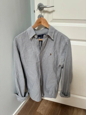 Grå skjorta från Ralph Lauren - Klassisk grå skjorta från Ralph Lauren med broderad logga på bröstet. Skjortan har långa ärmar, knappar framtill och button-down krage. Tillverkad i mjuk bomull som känns skön mot huden. Perfekt till jeans eller chinos. Vid frågor hör gärna av er!