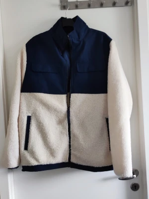 Blå och vit teddyjacka från 157 - Säljer en snygg teddyjacka från 157 i storlek M. Jackan har marinblå ovandel och detaljer samt krämvit, fluffig teddy på ärmar och nederdel. Dragkedja framtill, ståkrage och två fickor med blå kant. Perfekt för kalla dagar och riktigt skön att ha på sig.