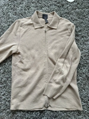 Beige zip-cardigan med krage - Stilren beige cardiganjacka med dragkedja framtill och klassisk krage. Tillverkad i mjukt, stickat material och har långa ärmar. Funkar som jacka och tröja! Tröjan är i väldigt bra skick!