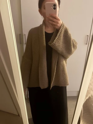 Beige oversized stickad kofta - Mysig oversized stickad kofta i beige med bred krage och knytskärp i midjan. Koftan har en avslappnad passform och grovstickad struktur, perfekt att slänga över en enkel outfit för extra stilpoäng.