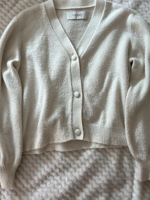Krämvit kofta från Studio Only - Mysig krämvit kofta från Studio Only med v-ringning och tre stora knappar framtill. Koftan har långa ärmar, ribbade muddar och en mjuk, lite fluffig struktur. Perfekt att slänga över en topp för en chill look.