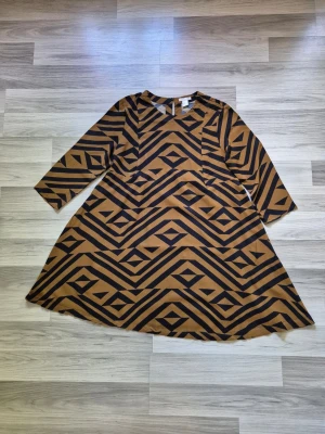 Brun och svart mönstrad klänning H&M - Cool klänning från H&M i brun och svart med grafiskt mönster. Klänningen har trekvartsärm och en A-linjeformad siluett som ger en avslappnad look. Tillverkad i mjuk bomull, perfekt för dig som gillar unika prints.