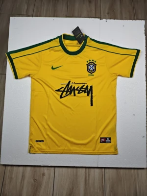 Gul Brasilien fotbollströja Stussy - Gul Brasilien fotbollströja från Nike med gröna detaljer och Stussy-logga på bröstet. Tröjan har rund hals, korta ärmar och Brasilien-emblem med fem stjärnor. Perfekt för dig som vill sticka ut på planen eller på läktaren.