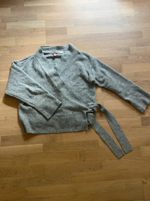 Grå omlottkofta Medium  - En grå kofta i omlottmodell och snörning i sidan.  Storlek M från H&M.  Material: 50% polyester, 29% polyamid, 13% akryl, 5% ull, 3% elestan