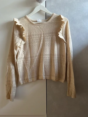 Beige stickad tröja med volang - Söt beige stickad tröja med volangdetaljer längs axlarna. Tröjan har ett genombrutet mönster, rund halsringning och långa ärmar. Perfekt för dig som gillar en romantisk och trendig stil. Materialet känns mjukt och lätt. Ett lite hål strax innan armhålan men det syns inte när man har på sig den.