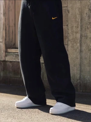 Nike x Nocta byxor - Säljer ett par svarta mjukisbyxor från Nike x Nocta. Avslappnad passform och gul broderad swoosh på vänster ben. Byxorna har dragsko i midjan och sidofickor. Perfekta för chill dagar eller streetwear-stil.