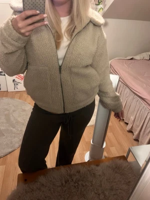 Beige teddyjacka med krage - Mysig beige teddyjacka med klassisk krage och dragkedja framtill. Jackan har två fickor och är gjord i fluffigt teddy-material som håller dig varm. Perfekt för dig som vill ha en trendig och bekväm look till hösten eller vintern.