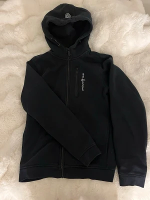 Svart zip hoodie från Sail Racing  - Säljer en svart hoodie från Sail Racing med dragkedja framtill och en bröstficka med vertikal zip. Hoodien har en broderad logga på huvan och tryckt logga på bröstet. Perfekt för dig som gillar enkel och sportig stil. Nypris 999kr🥰